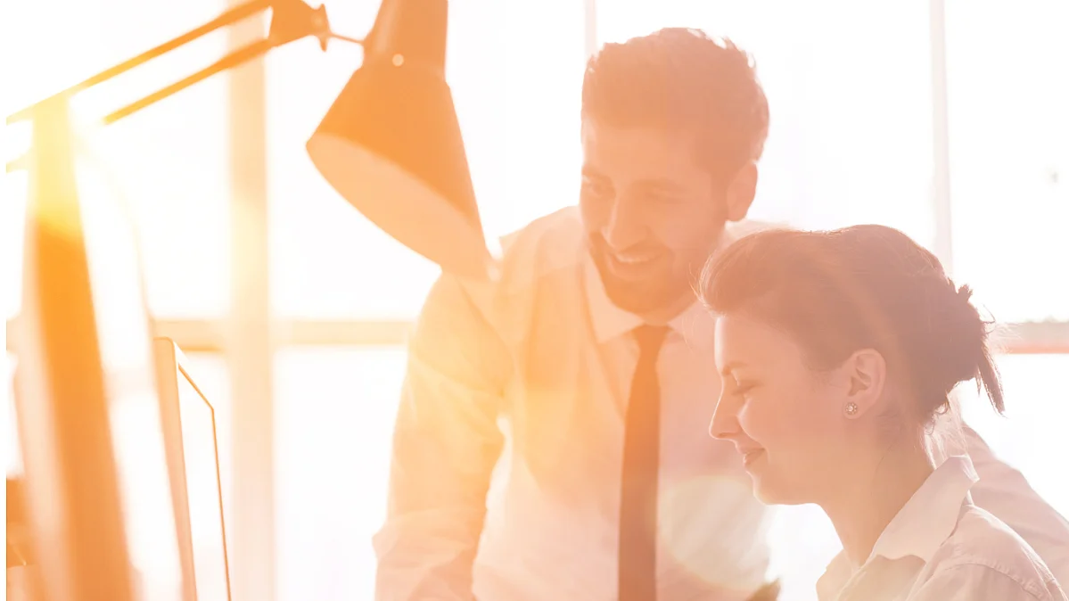 Eine Frau und ein Mann in Bürokleidung befinden sich vor einem großen Fenster, durch das die Sonne stark scheint. Eine Frau stizt und eine Frau Büroangestellter und eine Büroangestellte 